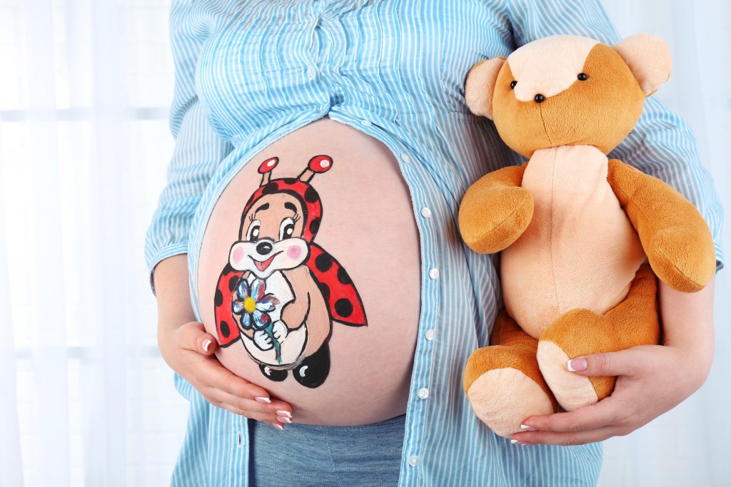 Beautiful young pregnant with baby toy  and picture on her belly, on light background